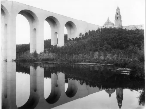 A large arched bridge spans over a river, reflecting in the water below, with a domed building and tower visible on a wooded hill in the background.