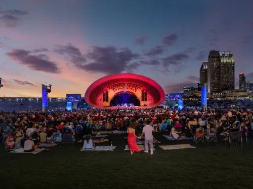 The Shell. A large crowd sits on a lawn facing an outdoor concert stage with a red-lit arch, city skyscrapers, and a sunset sky in the background.