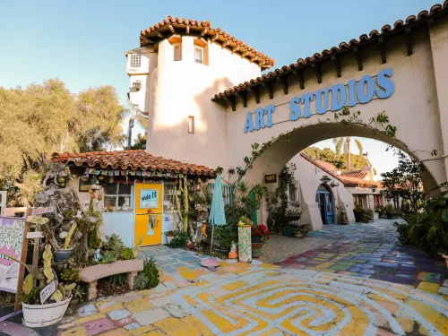 Colorful outdoor art studio with painted walkway, potted plants, tiled roofs, and a large “ART STUDIOS” sign above an archway on a sunny day.