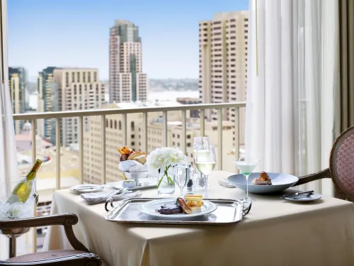 A table set for two at San Diego's Westgate Hotel, with gourmet meals, wine, and flowers by a window overlooking a city skyline with tall buildings under a clear sky.