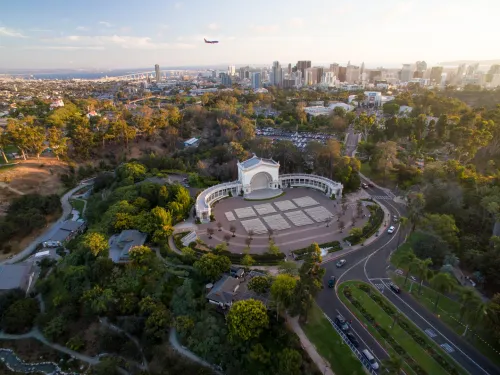 Aerial view of Balboa Park’s Spreckels Organ Pavilion in San Diego, with city skyline and a plane flying overhead in the background.