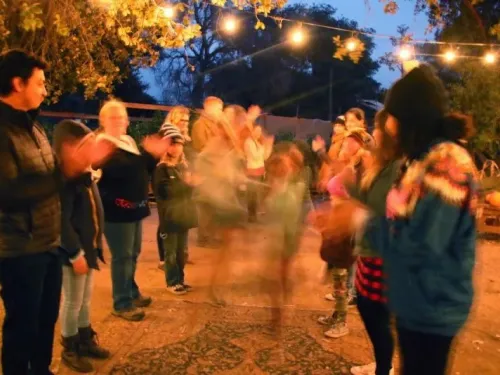 A group of people stands outdoors in a circle under string lights at dusk, some clapping and others blurred in motion, at the Fort Cross Haunted Harvest in Julian near San Diego.