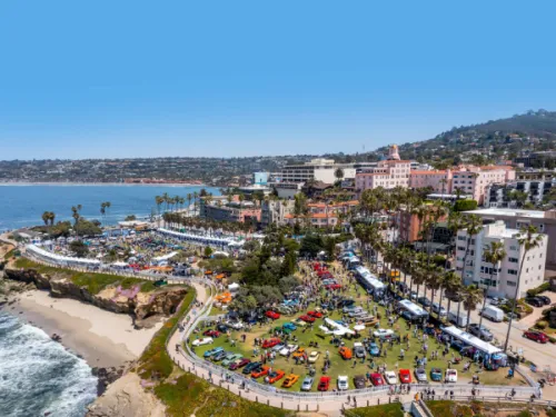 Aerial view of a coastal city park hosting a classic car show, with colorful cars on display, nearby buildings, palm trees, and the ocean in the background.