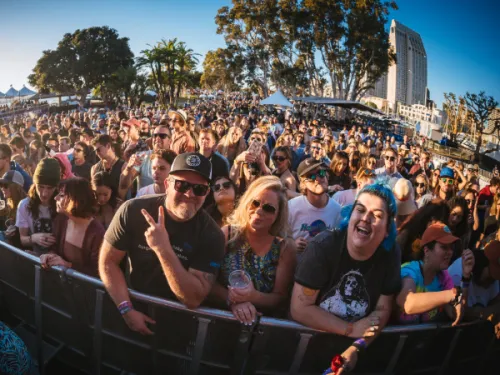 A large crowd gathers outdoors at a music festival in front of a stage on a sunny day, with people smiling and enjoying the event.