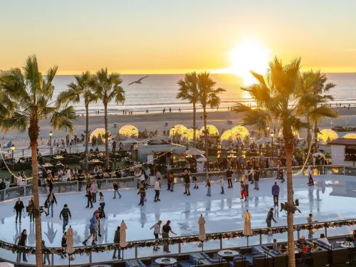 People skate on an outdoor ice rink by the beach at sunset, surrounded by palm trees, with the ocean and a clear sky in the background.