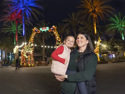 A woman holds a smiling child at night in front of palm trees and giraffe-shaped light displays, with colorful lights illuminating the background.