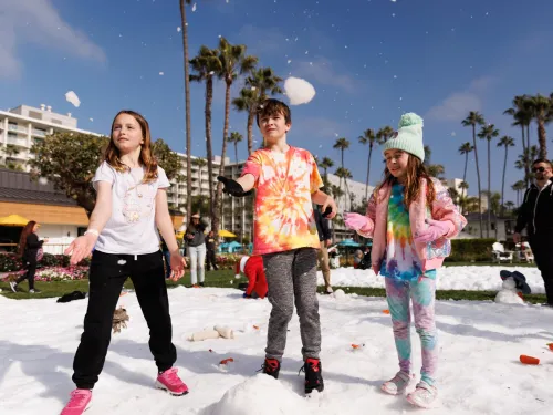 Three children in colorful clothes play with snow outdoors on a sunny day, with palm trees and buildings in the background at the Town and County Hotel in San Diego's Hotel Circle