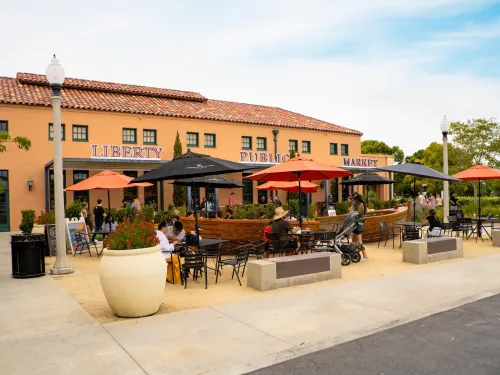 Outdoor seating area with tables, umbrellas, and people in front of the Liberty Public Market building on a clear day.