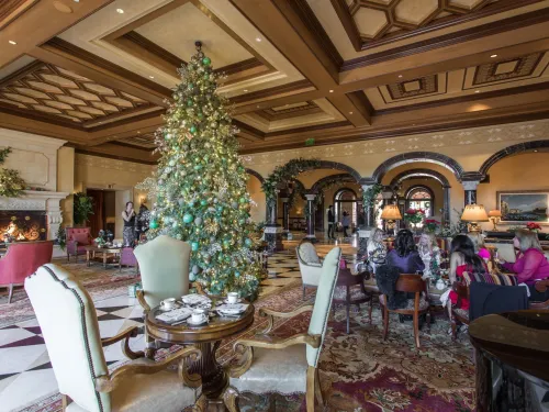 Decorated Christmas tree in a grand, ornate hotel lobby with guests seated at tables having tea near a lit fireplace.