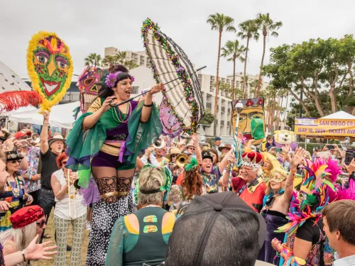 A performer on stilts holds a decorated umbrella above a lively, costumed crowd at an outdoor festival with masks, beads, and banners.