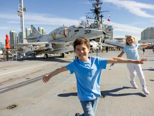 Two children in blue shirts pose with arms outstretched on the deck of the USS Midway Museum aircraft carrier, with a Navy jet and city buildings in the background.