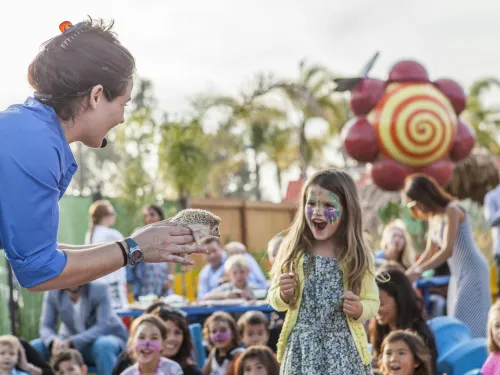 A woman shows a hedgehog to a smiling girl with face paint at an outdoor event with children and adults in the background.