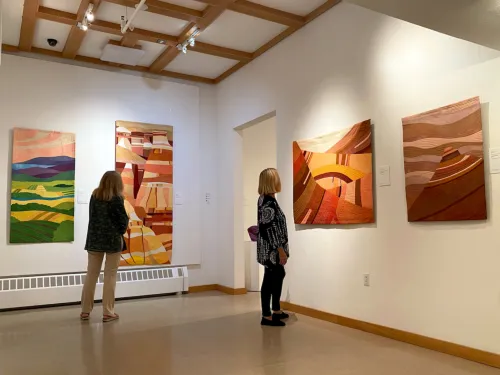 Two women view colorful abstract textile art pieces displayed on white walls in a well-lit gallery space of the Vision Museum of Textile Art