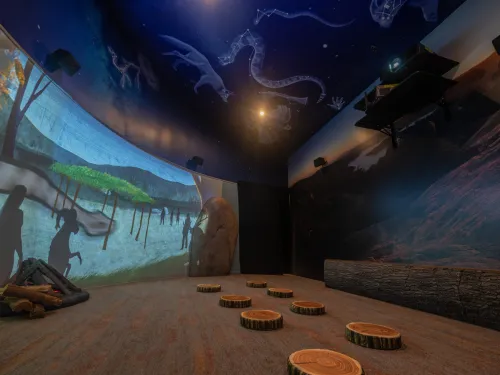 A dark room with projected images of constellations on the ceiling, nature scenes on the walls, and wooden stumps arranged on the carpeted floor at the Barona Cultural Center