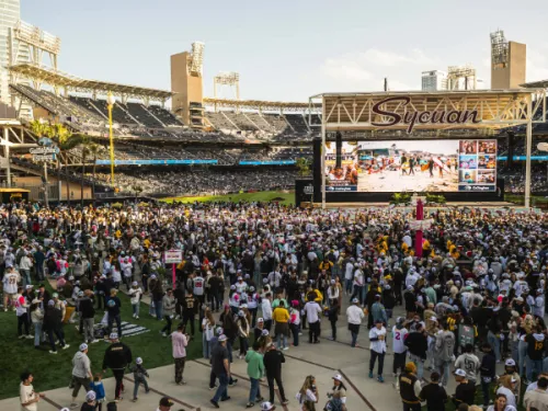 A large crowd gathers on a baseball field in front of a big screen showing images, with stadium seating and buildings in the background.