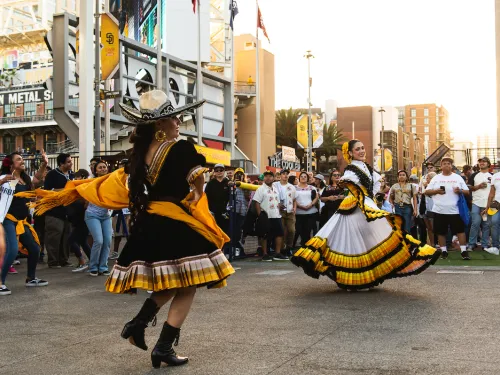 Two folklórico dancers perform in front of a crowd outside Petco Park in San Diego. The dancer in the foreground wears a black dress with yellow accents and a wide-brimmed sombrero, twirling a yellow shawl, while the dancer behind her wears a flowing white, yellow, and black ruffled dress, holding out the skirt as she spins. Baseball fans in jerseys and caps watch from behind a barrier, with stadium signage, flags, and downtown buildings visible in the background at sunset.