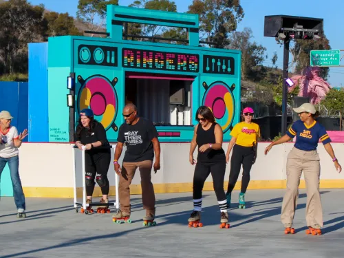 Five adults roller skating side by side at an outdoor rink at Ringers Roller Rink in San Diego’s Encanto neighborhood, all smiling and mid-glide in the sun. Behind them is a brightly colored backdrop designed like a giant boombox with the word “Ringers” on it, framed by trees and blue sky.