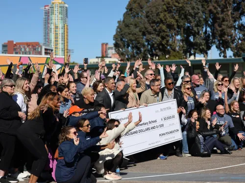 Large group of adults outdoors at a corporate volunteer or charity event, smiling and raising their hands as they pose together around two oversized donation checks held at the center of the group, with city buildings and trees in the background.
