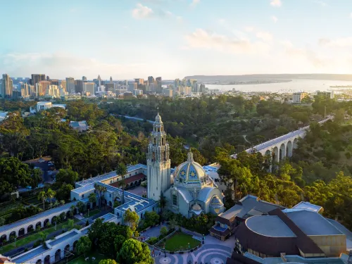 Elevated view of Balboa Park featuring the California Tower and historic buildings amid lush greenery, with the San Diego skyline stretching across the horizon and San Diego Bay beyond.