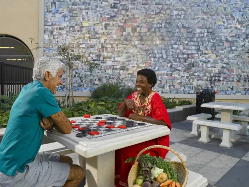 Two people sit across from each other playing a game with large red and black checkers on a stone table in an outdoor plaza in City Heights, San Diego, California. The person on the left wears a teal shirt and khaki shorts; the person on the right wears a bright red dress with gold embroidery and is smiling. A large mosaic-style mural made of many small photos fills the wall behind them, and a basket of fresh vegetables and herbs rests on the ground beside the table.