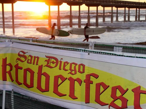 Sunset at the Ocean Beach Pier with surfers in the background and a banner promoting Ocean Beach Oktoberfest in San Diego.
