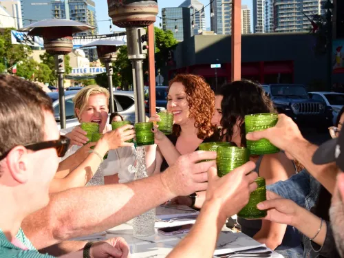 A group of people seated at an outdoor restaurant table raise their glasses in a toast under sunny skies in San Diego's Little Italy neighborhood.