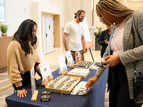 A Tourism Accelerator member stands behind a display table at the San Diego Convention Center, showcasing neatly arranged products in trays and stands. Informational signs with photos and branding are propped up on the table. A visitor in the foreground examines one of the items, while another person stands nearby in the background. The setting appears to be an indoor expo or networking event.