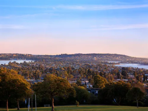 Wide panoramic view from Kate Sessions Park in Pacific Beach, San Diego, showing rolling green hills, trees, and the city and coastline at sunset.