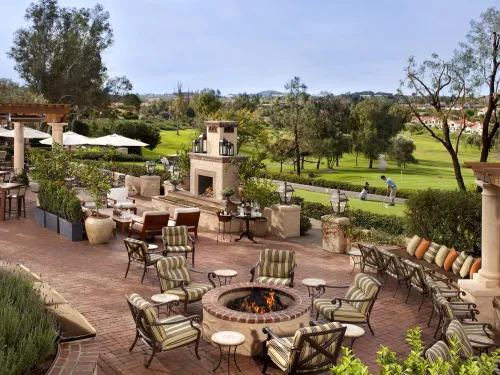 A daytime view of an outdoor patio at the Rancho Bernardo Inn in San Diego, with fire pits and furniture and a golf course beyond.