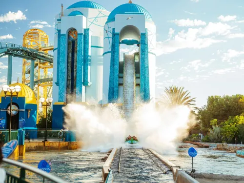 Splashdown moment on the Journey to Atlantis ride at SeaWorld San Diego, with a boat racing down the final drop and water erupting in front of the iconic blue and white Atlantis towers under a sunny sky.
