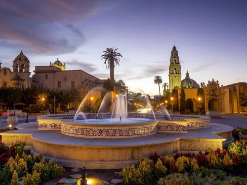 Balboa Park Fountain surrounded by vibrant flowers at dusk, with historic buildings and a clock tower in the background.