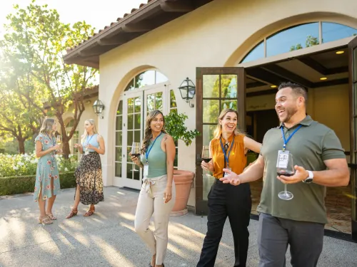 Small group networking outdoors at a San Diego venue, with attendees holding drinks and chatting near a Spanish-style building surrounded by trees and warm afternoon light.