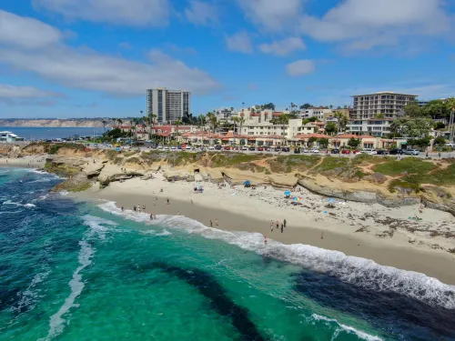 Aerial view of La Jolla Cove in San Diego, showing turquoise ocean waters, a sandy beach with visitors, rugged coastal cliffs, and nearby buildings under a bright blue sky.