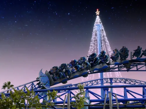 Nighttime ride on the Manta roller coaster at SeaWorld San Diego during the Christmas Celebration, with the train soaring along blue track in front of a towering, illuminated holiday tree against a twilight sky.