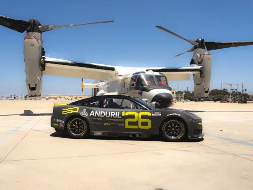 A sleek black NASCAR racing car with the number 26 is parked in front of a U.S. Navy Osprey aircraft on a sunny day at Naval Base Coronado near San Diego.