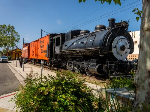 Vintage steam locomotive next to an orange boxcar in a sunny outdoor setting.