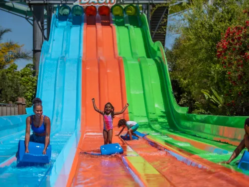 Children sliding down colorful water slides on a sunny day, with vibrant greenery in the background.