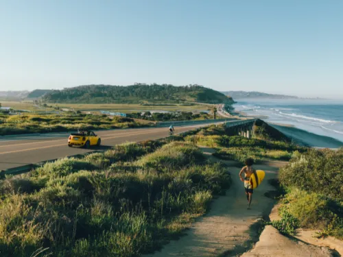 A person walks along a path by the shore, with waves and a coastline view.