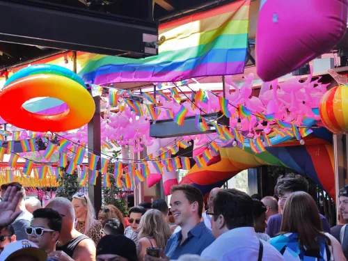 Colorful decorations and a crowd celebrate Pride, with rainbow flags and balloons.