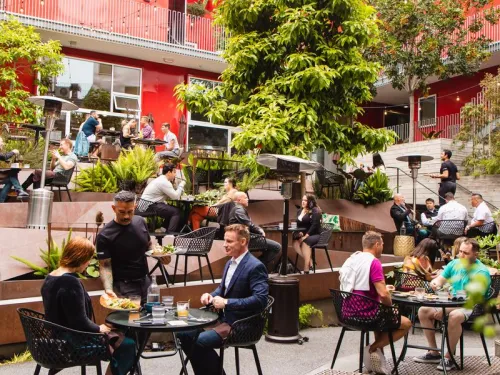 A lively outdoor café scene with people dining and socializing among greenery and colorful architecture.