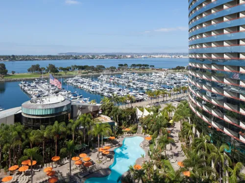 A waterfront view featuring a marina, a hotel, and a pool surrounded by palm trees.