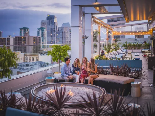 Group of friends chatting around a fire pit on a rooftop with the San Diego skyline in the background.