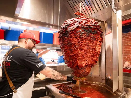 A chef slices meat from a large vertical rotisserie in a vibrant kitchen setting.