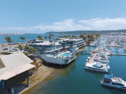 Aerial view of a marina with boats, buildings, and palm trees under a sunny sky.