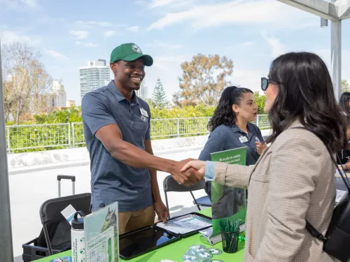 A man shakes hands with a woman at an information table outdoors at the career fair in San Diego at San Diego City College, with city buildings in the background.