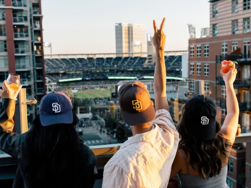 Three friends in baseball caps raise drinks at Borrego, overlooking a Petco Park at sunset.