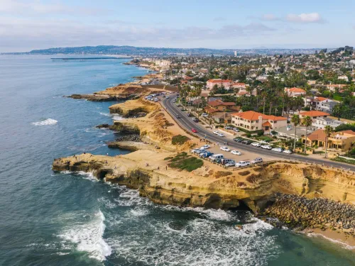Aerial view of the Sunset Cliffs coastal area with houses, cliffs, and crashing waves under a blue sky.