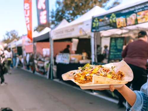 A hand holding a tray of tacos in a busy outdoor market at the Hillcrest Farmers Market in San Diego, with food stalls.