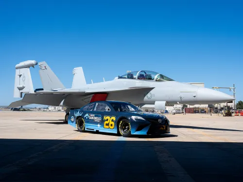 A blue and yellow race car parked beside a white fighter jet on a clear day.
