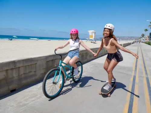 Two girls smile and hold hands while biking and skateboarding along the Mission Beach Boardwalk in San Diego.